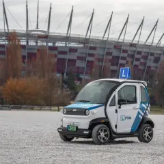 Mały biały samochód z niebieskimi pasami i literą "L" na dachu, na tle stadionu.