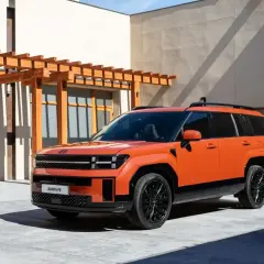 Pomarańczowy, tani SUV Hyundai Santa Fe z czarnymi felgami stoi przed budynkiem z drewnianą pergolą.