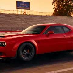 Czerwony Dodge Challenger SRT pędzi po torze wyścigowym podczas zachodu słońca, z trybunami w tle.
