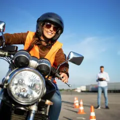 Uśmiechnięta kursantka w kasku i kamizelce na motocyklu, gotowa do jazdy. Instruktor obserwuje ją na placu manewrowym. kategoria a.