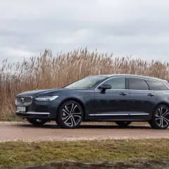 Ciemnogranatowe Volvo v90 na tle wysokiej, suchej trawy i pochmurnego nieba.
