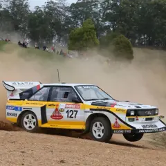 Klasyczny Audi Quattro z grupy B, oznaczony numerem 127, pędzi po szutrowej drodze, wzbijając tumany kurzu.