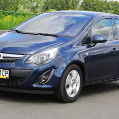 Niebieski Opel Corsa D 1.4 benzyna w ruchu drogowym, z zielenią drzew w tle.