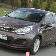 Brązowe Kia Rio 3 w ruchu drogowym, z widocznym grillem i reflektorami.