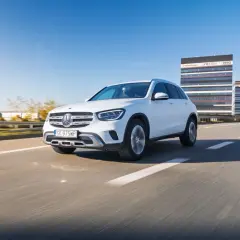 Biały Mercedes GLC 200 jedzie autostradą w słoneczny dzień, z nowoczesnymi budynkami w tle.