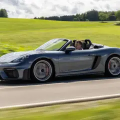 Szary Porsche Spyder RS z kierowcą pędzi po wiejskiej drodze, otoczony zielonymi wzgórzami.