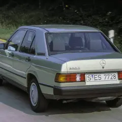 Klasyczny zielony Mercedes 190E z lat 80. sunie po drodze wśród drzew.
