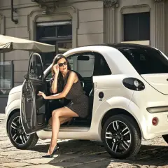 Elegancka kobieta w okularach przeciwsłonecznych wysiada z białego samochodu Microcar. Idealny samochód bez prawa jazdy na miejskie wypady.