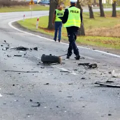 Na drodze po kolizji leżą szczątki pojazdów i opona. Policjanci w kamizelkach odblaskowych badają miejsce zdarzenia, być może szukając śladów ucieczki z miejsca kolizji.