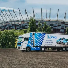 Ciężarówka z logo "Linia Szkolenia Kierowców" i samochodami na naczepie, na tle Stadionu Narodowego. Marzysz o **prawie jazdy na tira**? Tu możesz je zdobyć!