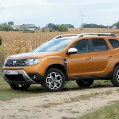 Pomarańczowy Dacia Duster 1.6 benzyna na polnej drodze, w tle pole kukurydzy i domy.