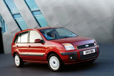 Czerwony Ford Fusion 1.4 benzyna w ruchu, z kobietą za kierownicą, na tle nowoczesnego budynku.