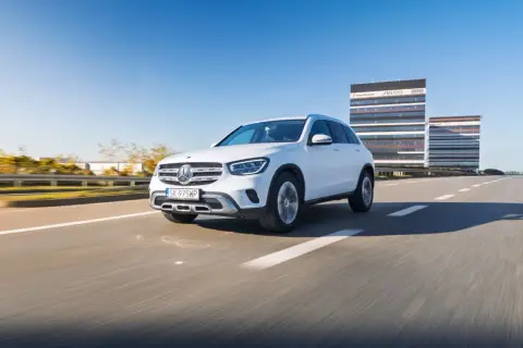 Biały Mercedes GLC 200 jedzie autostradą w słoneczny dzień, z nowoczesnymi budynkami w tle.