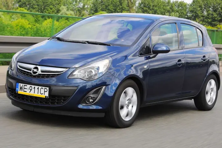 Niebieski Opel Corsa D 1.4 benzyna w ruchu drogowym, z zielenią drzew w tle.