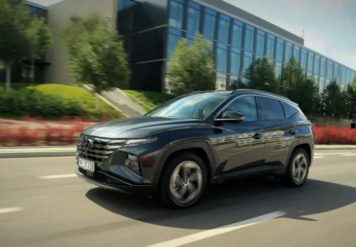 Ciemnoszary Hyundai Tucson 2024 pędzi po drodze, mijając nowoczesny budynek i zielone drzewa.