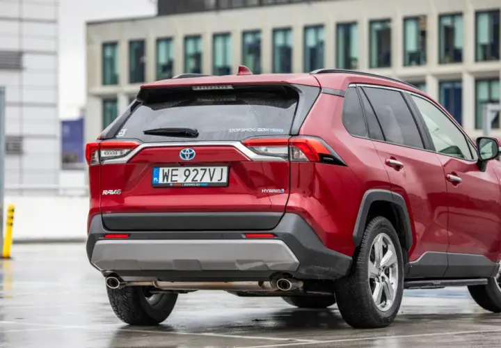 Czerwony, używany Toyota RAV4 Hybrid z salonu, zaparkowany na mokrym parkingu przed nowoczesnym budynkiem.