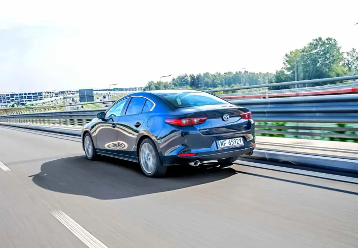 Ciemnoniebieska Mazda 3 sedan jedzie po autostradzie, odbijając światła i otoczenie.