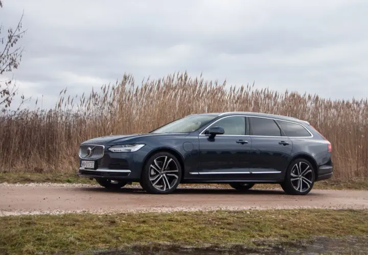 Ciemnogranatowe Volvo v90 na tle wysokiej, suchej trawy i pochmurnego nieba.