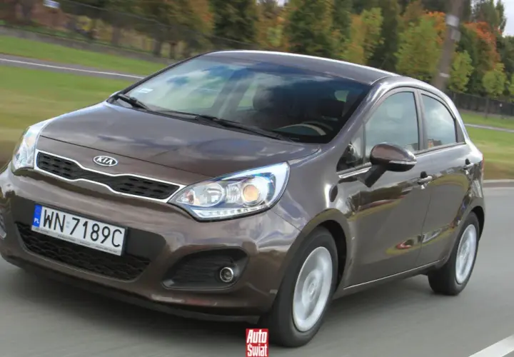 Brązowe Kia Rio 3 w ruchu drogowym, z widocznym grillem i reflektorami.
