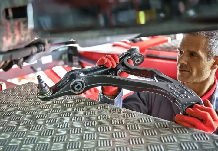 Mechanik w czerwonych rękawiczkach prezentuje element zawieszenia samochodu.