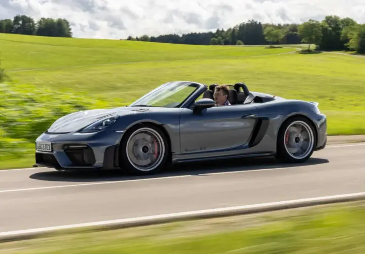 Szary Porsche Spyder RS z kierowcą pędzi po wiejskiej drodze, otoczony zielonymi wzgórzami.