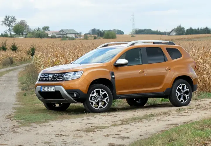 Pomarańczowy Dacia Duster 1.6 benzyna na polnej drodze, w tle pole kukurydzy i domy.