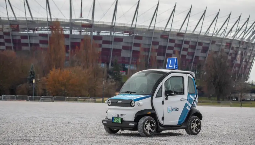 Mały biały samochód z niebieskimi pasami i literą "L" na dachu, na tle stadionu.