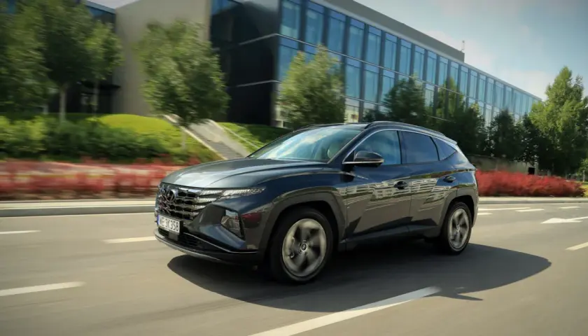 Ciemnoszary Hyundai Tucson 2024 pędzi po drodze, mijając nowoczesny budynek i zielone drzewa.