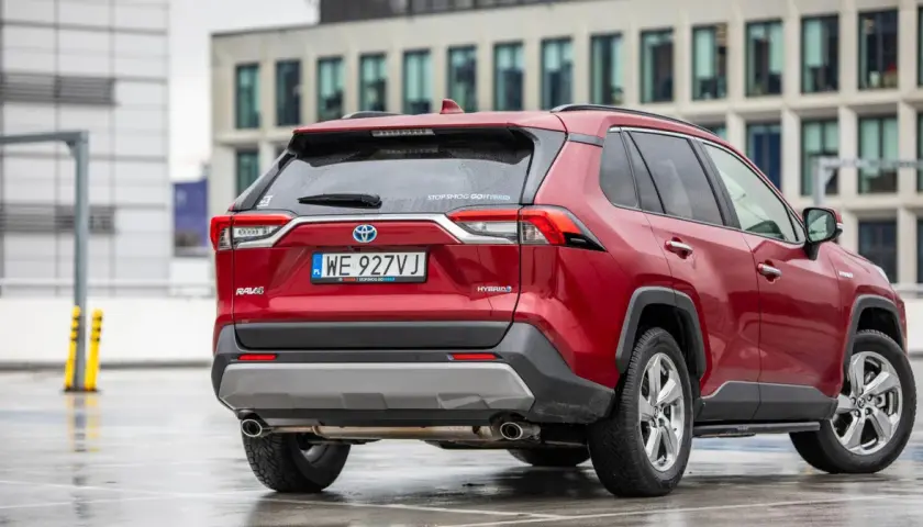 Czerwony, używany Toyota RAV4 Hybrid z salonu, zaparkowany na mokrym parkingu przed nowoczesnym budynkiem.
