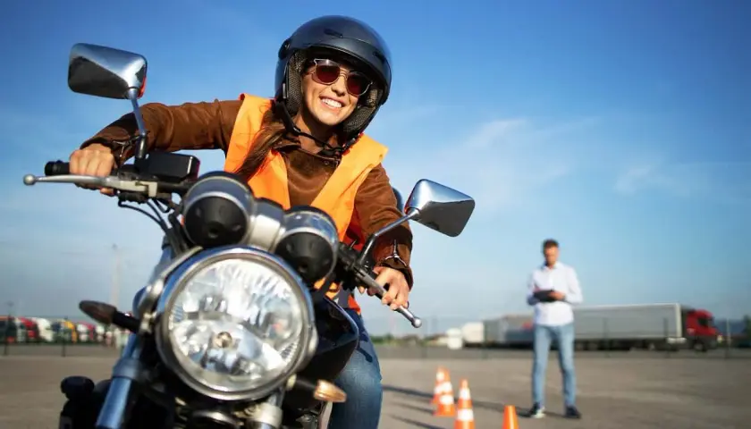 Uśmiechnięta kursantka w kasku i kamizelce na motocyklu, gotowa do jazdy. Instruktor obserwuje ją na placu manewrowym. kategoria a.
