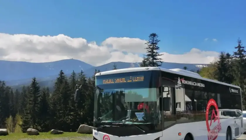 Biały autobus "Karpacz Bus" z napisem "Komunikacja Miejska" na tle gór i lasu. Zastanawiasz się, ile kosztuje autobus?