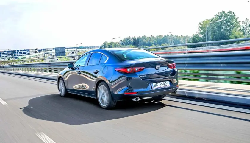 Ciemnoniebieska Mazda 3 sedan jedzie po autostradzie, odbijając światła i otoczenie.
