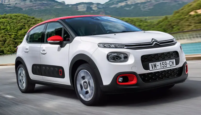 Biały Citroen C3 2025 z czerwonym dachem i lusterkami jedzie po drodze, otoczony zielonymi wzgórzami.
