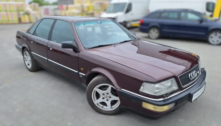 Bordowy Audi V8 z lat 90. na parkingu, w tle białe vany i niebieski kombi.