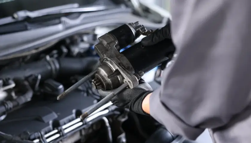 Mechanik w czarnych rękawiczkach trzyma rozrusznik w aucie, gotowy do montażu.