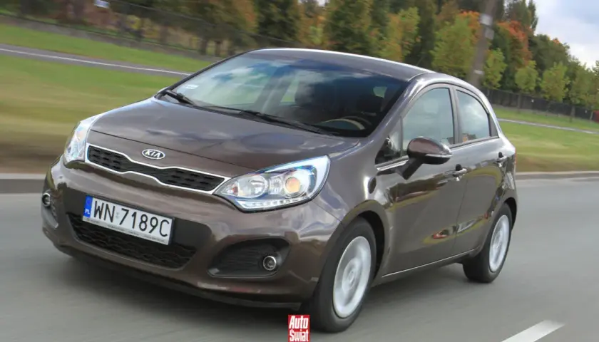 Brązowe Kia Rio 3 w ruchu drogowym, z widocznym grillem i reflektorami.