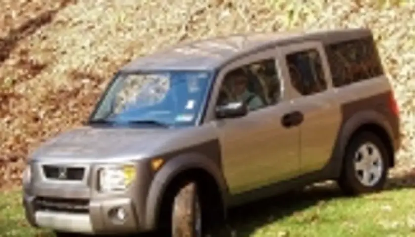 Srebrny Honda Element z brązowymi elementami, zaparkowany na trawie przy zboczu pokrytym liśćmi.