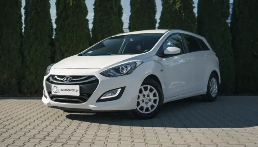 Biały Hyundai i30 1.4 benzyna na tle zielonych drzew, zaparkowany na kostce brukowej.