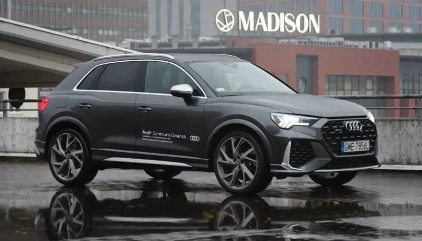 Szary Audi RS Q3 na mokrej nawierzchni, z odbiciem w kałuży. W tle budynek z napisem MADISON.