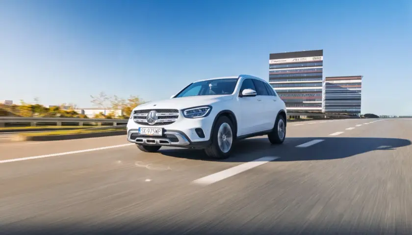 Biały Mercedes GLC 200 jedzie autostradą w słoneczny dzień, z nowoczesnymi budynkami w tle.