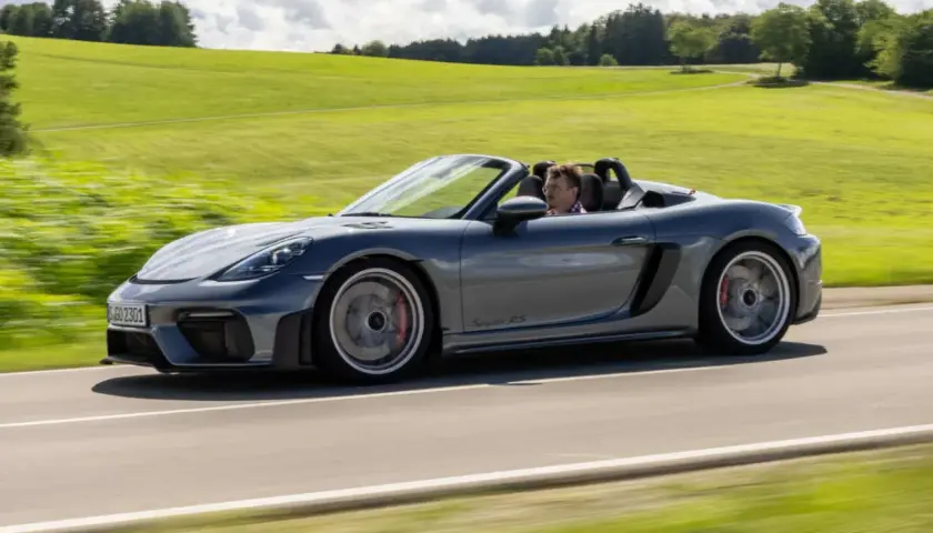 Szary Porsche Spyder RS z kierowcą pędzi po wiejskiej drodze, otoczony zielonymi wzgórzami.