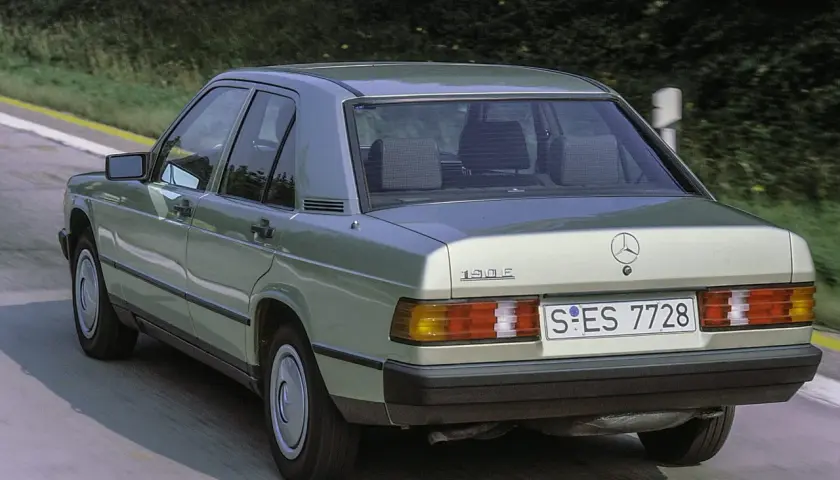Klasyczny zielony Mercedes 190E z lat 80. sunie po drodze wśród drzew.