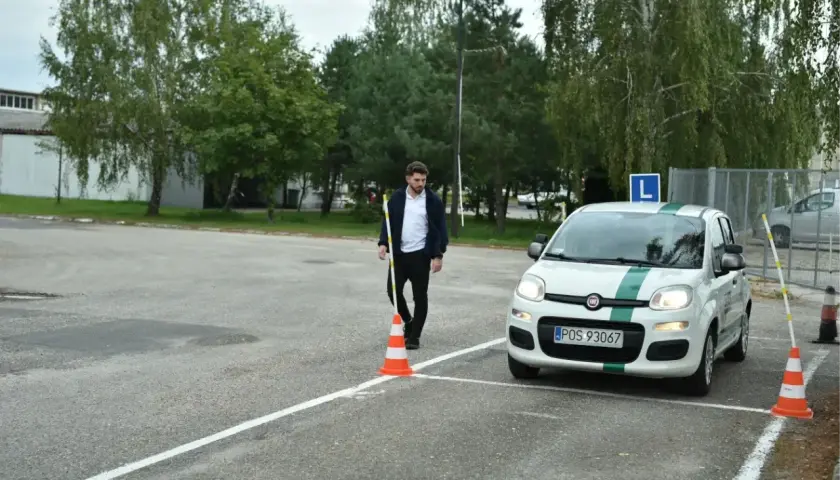 Młody mężczyzna ustawia pachołek na placu manewrowym. Obok stoi biały Fiat Panda z literą "L" na dachu, gotowy do egzaminu.