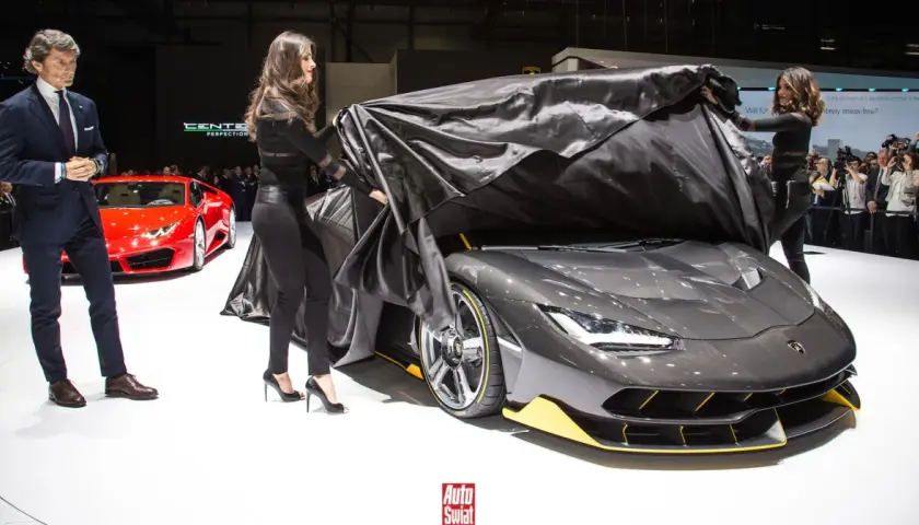 Elegancki mężczyzna i dwie kobiety odsłaniają nowy, szary Lamborghini Centenario. Obok stoi czerwony sportowy samochód.