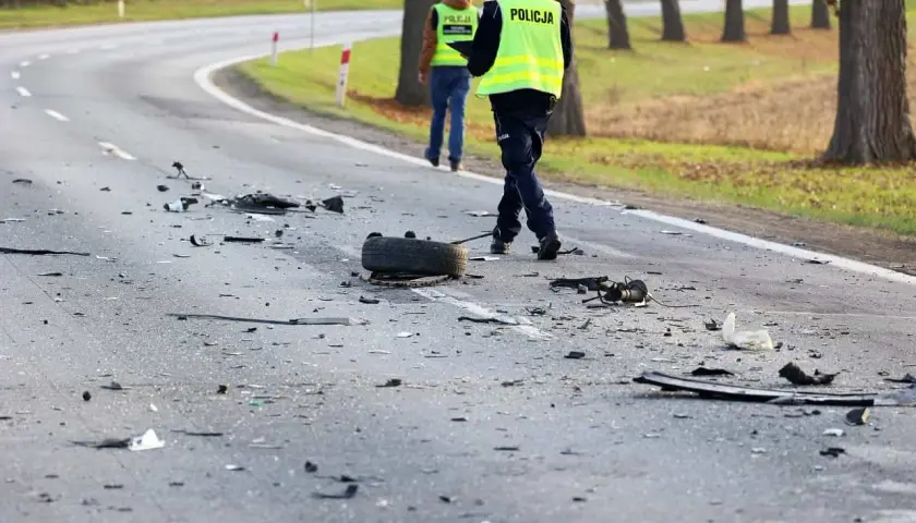 Na drodze po kolizji leżą szczątki pojazdów i opona. Policjanci w kamizelkach odblaskowych badają miejsce zdarzenia, być może szukając śladów ucieczki z miejsca kolizji.