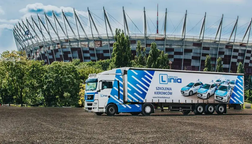 Ciężarówka z logo "Linia Szkolenia Kierowców" i samochodami na naczepie, na tle Stadionu Narodowego. Marzysz o **prawie jazdy na tira**? Tu możesz je zdobyć!
