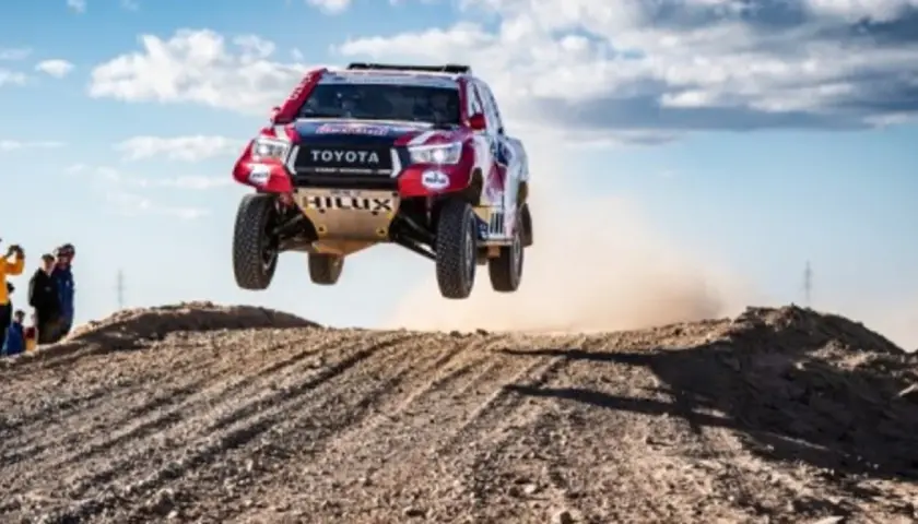 Toyota Hilux w locie podczas rajdu Dakar. Widzowie obserwują zmagania na pustynnym terenie.
