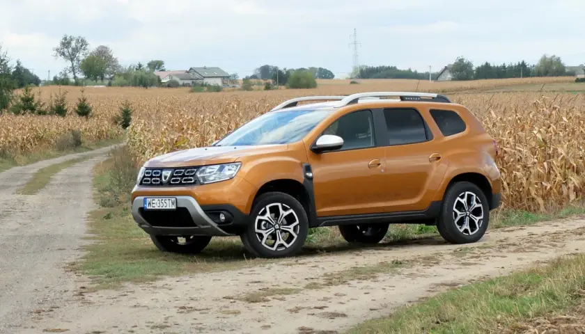 Pomarańczowy Dacia Duster 1.6 benzyna na polnej drodze, w tle pole kukurydzy i domy.