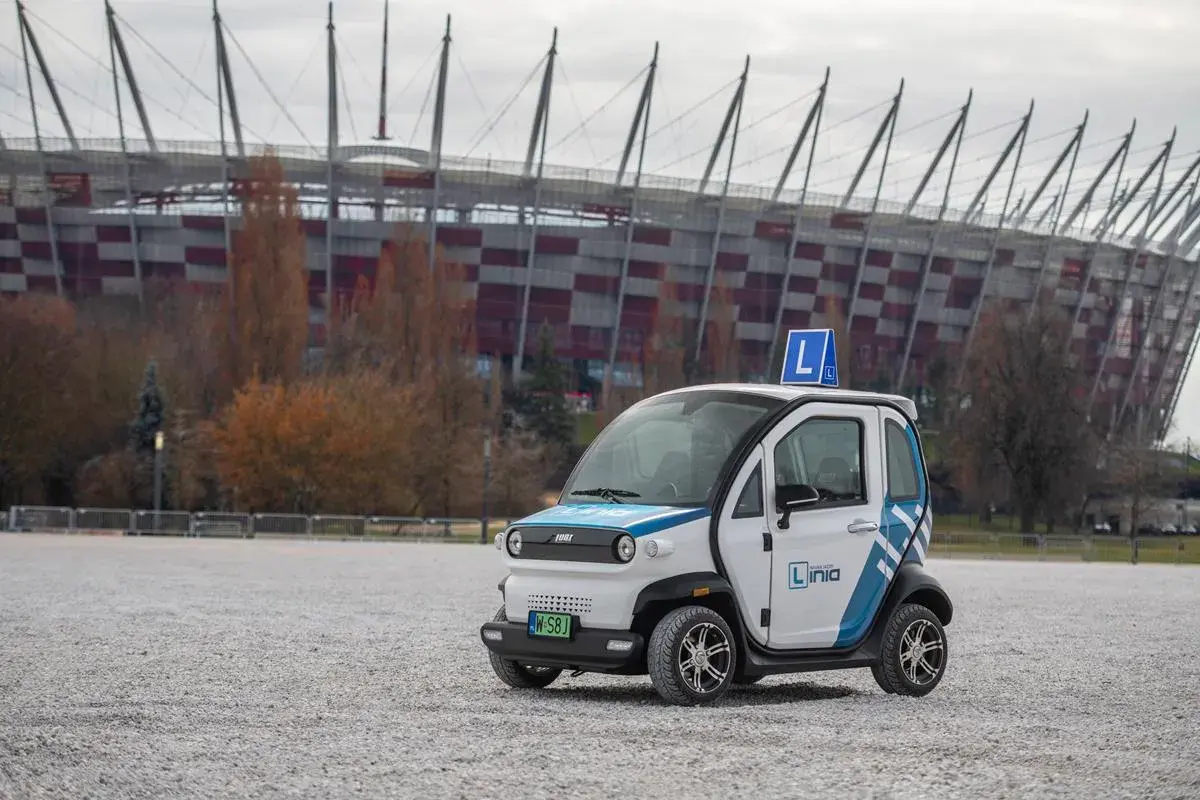 Mały biały samochód z niebieskimi pasami i literą "L" na dachu, na tle stadionu.