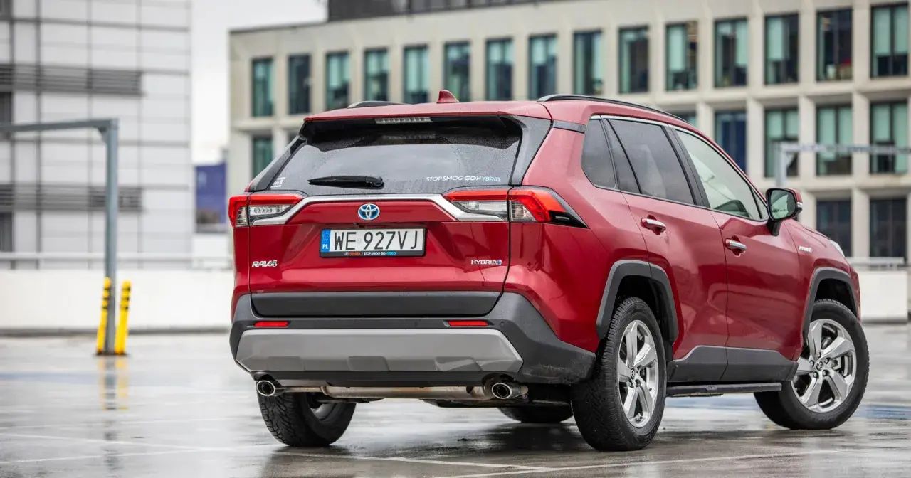 Czerwony, używany Toyota RAV4 Hybrid z salonu, zaparkowany na mokrym parkingu przed nowoczesnym budynkiem.
